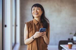 Woman standing indoors, smiling while holding a smartphone. She is wearing a sleeveless, button-up top and a wristwatch