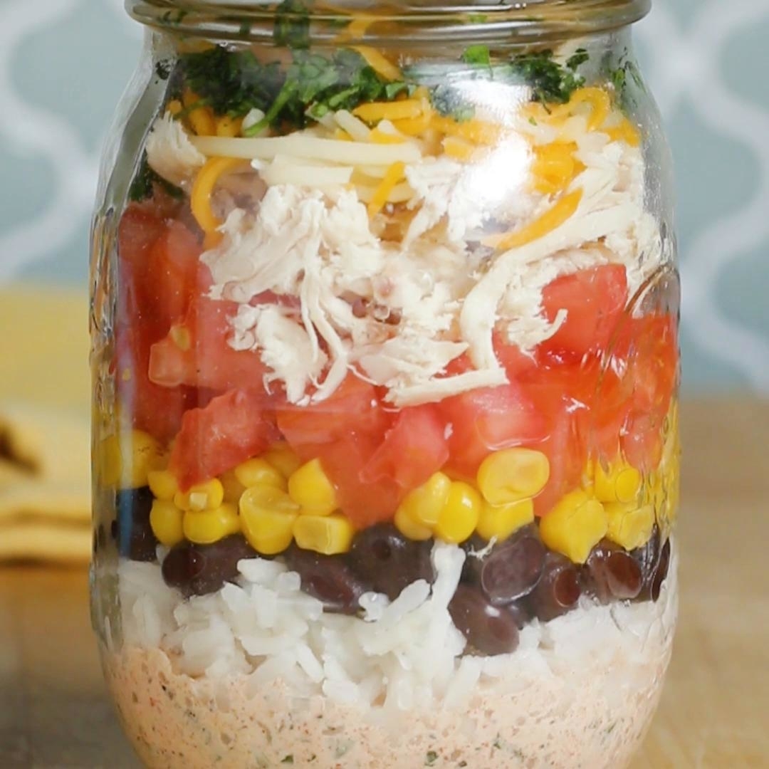 A mason jar filled with layered ingredients including rice, black beans, corn, diced tomatoes, shredded chicken, cheese, and herbs