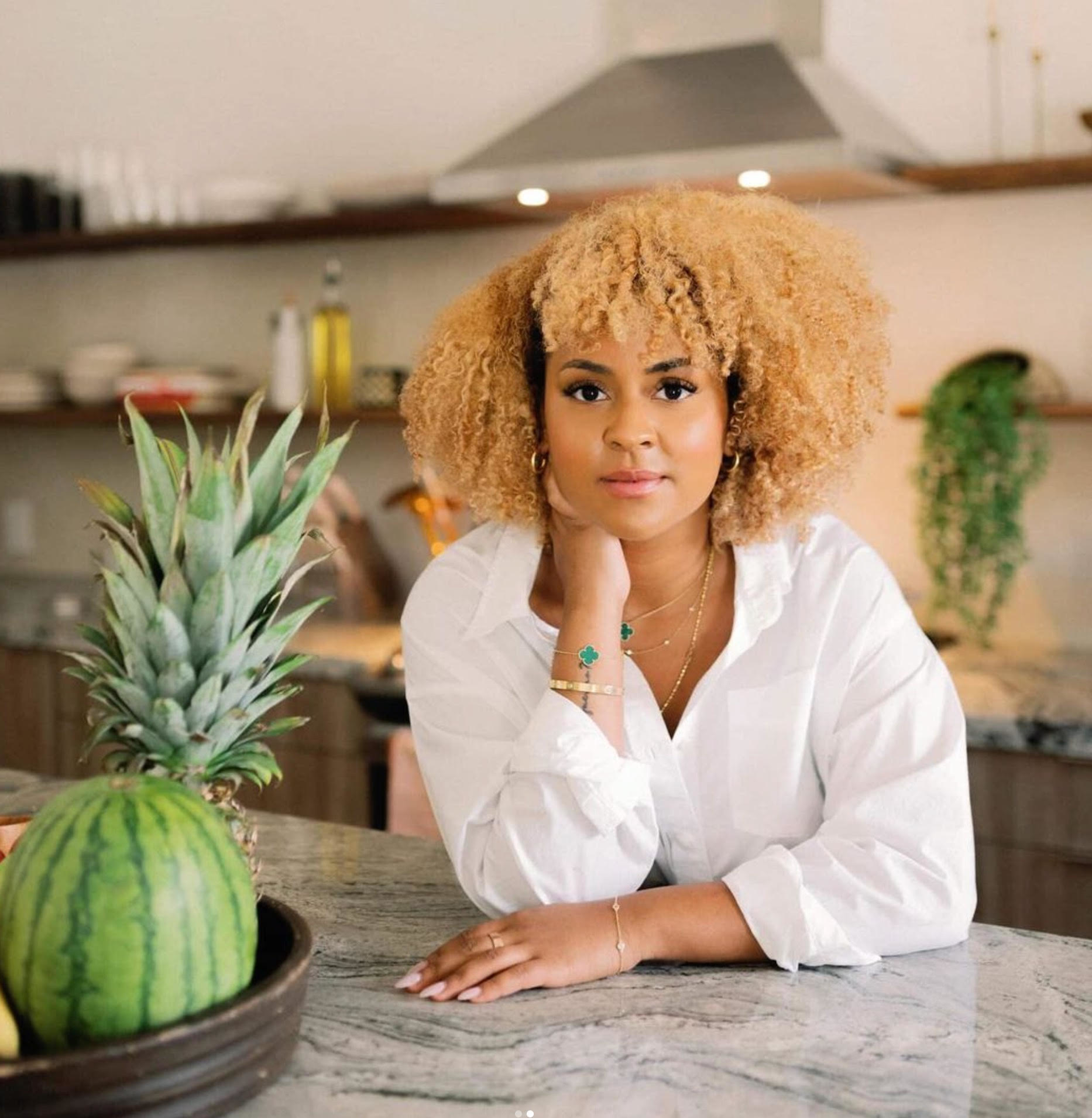 A woman with curly hair, dressed in a white blouse, leans on a kitchen counter adorned with a pineapple and watermelon in the foreground