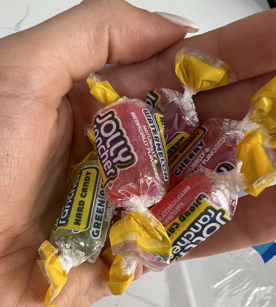 A hand holds a variety of Jolly Rancher hard candies in their wrappers, including watermelon, cherry, and green apple flavors